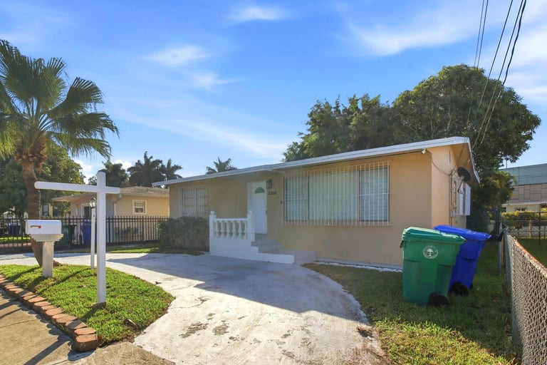 Yellow and white single-story residential home with metal roof, driveway, and trash cans in front, surrounded by palm trees and green lawn