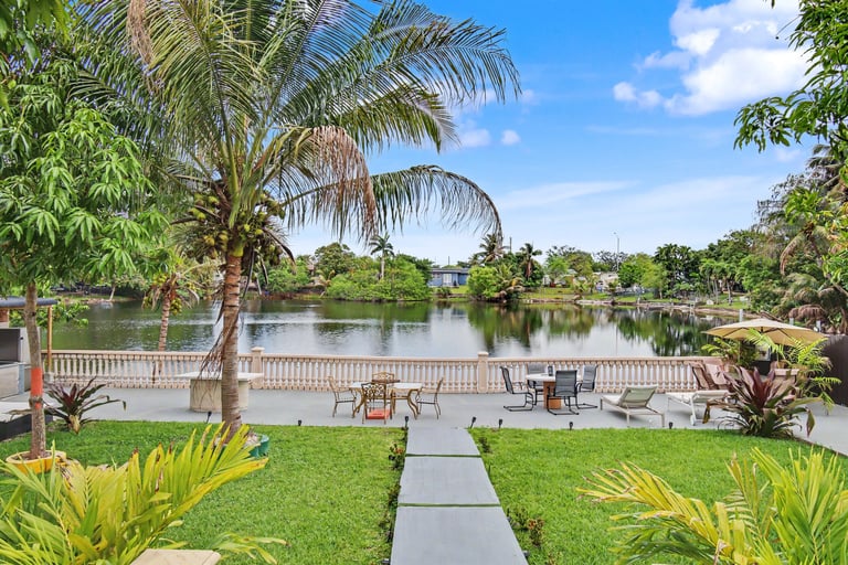 Tropical backyard with manicured lawn, palm trees, and waterfront view of a calm river or lake with dock and seating areas.