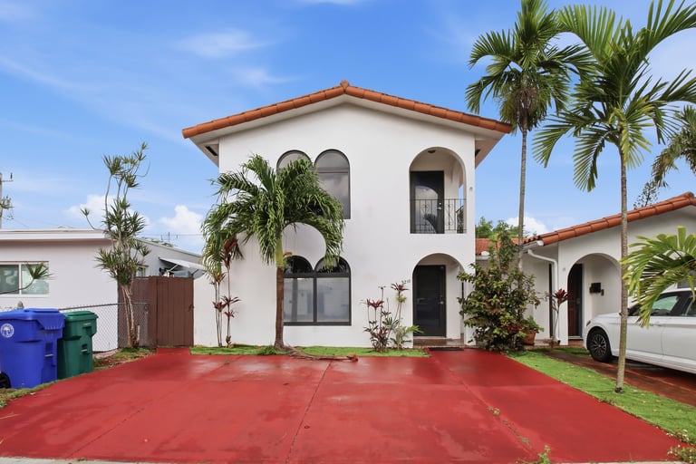 White two-story Spanish colonial house with terracotta roof and red driveway, palm trees, and attached garage under blue sky